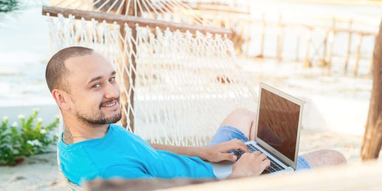 A man works on a laptop while lying in a hammock on the beach.