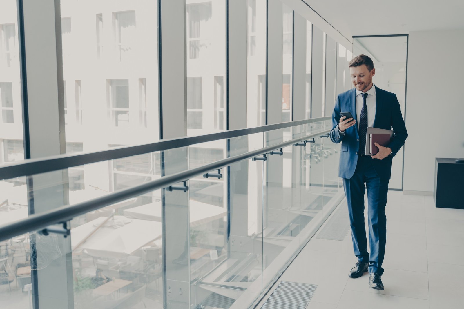 Smiling redhead man office worker in suit using cellphone while standing in modern office