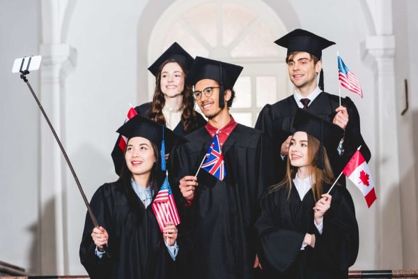 cheerful students in graduation gowns holding flags of different countries and taking selfie on